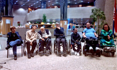 Skupina umjetnika i sudionika, od kojih su neki u invalidskim kolicima, sjedi u polukrugu u prostranom predvorju javne zgrade. Osobe međusobno razgovaraju i slušaju, a u pozadini se vide stupovi, rasvjeta i zastava Republike Hrvatske, tijekom događanja vezanog uz izložbu umjetnika koji slikaju nogama i ustima.