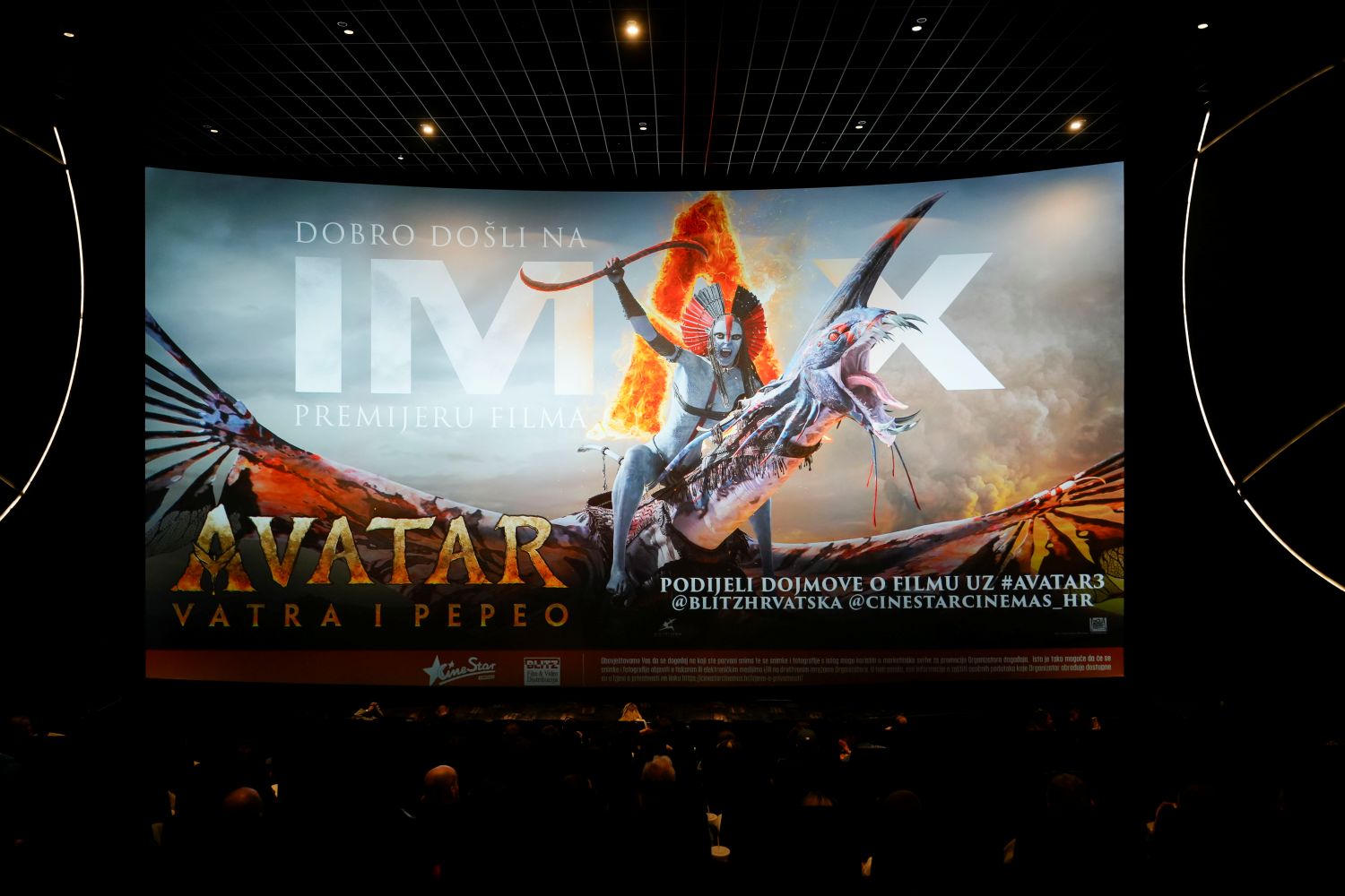 Fotografija prikazuje IMAX kino dvoranu tijekom premijere filma „Avatar: Vatra i Pepeo”. Na velikom kino platnu dominira promotivni plakat filma s dramatičnim prizorom: lik jaše leteće stvorenje, drži oružje i okružen je plamenom, što stvara snažan vizualni dojam akcije i fantazije. U dvorani su gledatelji već smješteni i gledaju prema platnu, što dodatno naglašava atmosferu iščekivanja i uzbuđenja. Na ekranu se nalazi i tekstualna dobrodošlica: „Dobro došli na IMAX premijeru filma Avatar: Vatra i Pepeo”, uz poziv da se dojmovi podijele na društvenim mrežama s oznakom #AVATAR3 te oznakama @BLITZHRVATSKA i @CINESTARCINEMAS_HR. U donjem dijelu ekrana istaknuti su logotipi CineStara i Blitz-a, a prisutna je i napomena da je kino opremljeno kamerama za snimanje i fotografiranje u promotivne svrhe, uz obavijest da ulaskom u prostor korisnici pristaju na takvo dokumentiranje.
