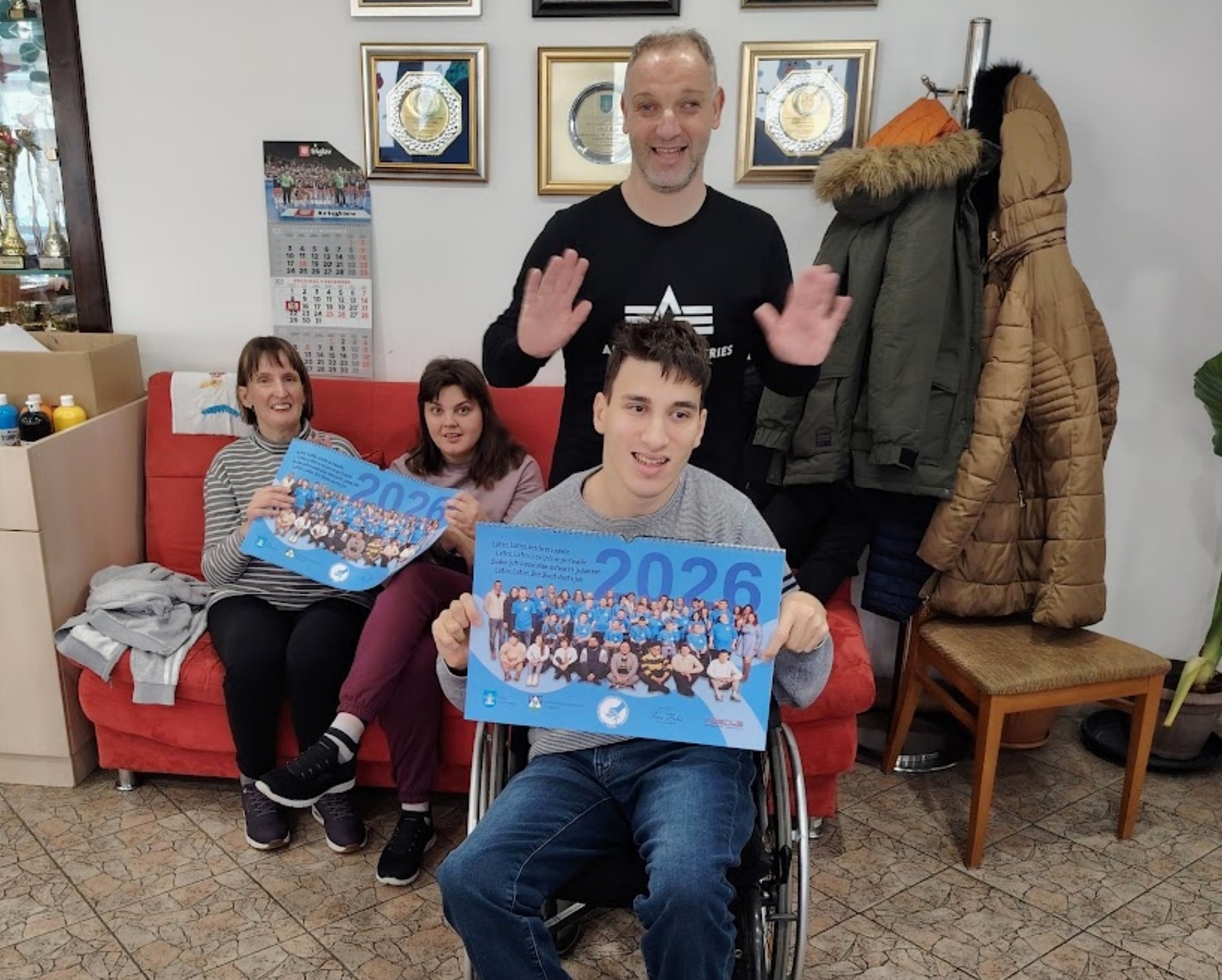 Na fotografiji se nalaze četiri osobe u zatvorenom prostoru, vjerojatno u zajedničkom prostoru poput društvenog centra ili udruge. Tri osobe sjede na crvenom kauču, dok jedna stoji iza njih s osmijehom i podignutim rukama u znak pozdrava. Osoba u prvom planu nalazi se u invalidskim kolicima i drži plakat ili kalendar s velikim plavim brojevima "2026". Dvije osobe pored nje također drže slične plakate. Na plakatima piše: "2026 Let's! Let's be in the circle Let's be in the circle of inclusion Let's be in the circle of friendship Let's be in the circle of love" U pozadini se vide kaputi na vješalici, kalendar za prosinac 2023., vitrina s trofejima i ormarić s bocama, što dodatno sugerira da se radi o prostoru za zajedničke aktivnosti. Atmosfera je topla i prijateljska, a poruka na plakatima naglašava inkluziju, prijateljstvo i ljubav — moguće u kontekstu kampanje, događanja ili inicijative za 2026. godinu.