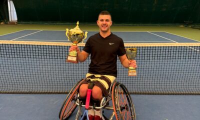 Slika prikazuje tenisača u kolicima kako drži dva pehara, a reket mu je naslonjen na noge. On se nalazi na teniskom terenu, a iza njega je vidljiva teniska mreža.