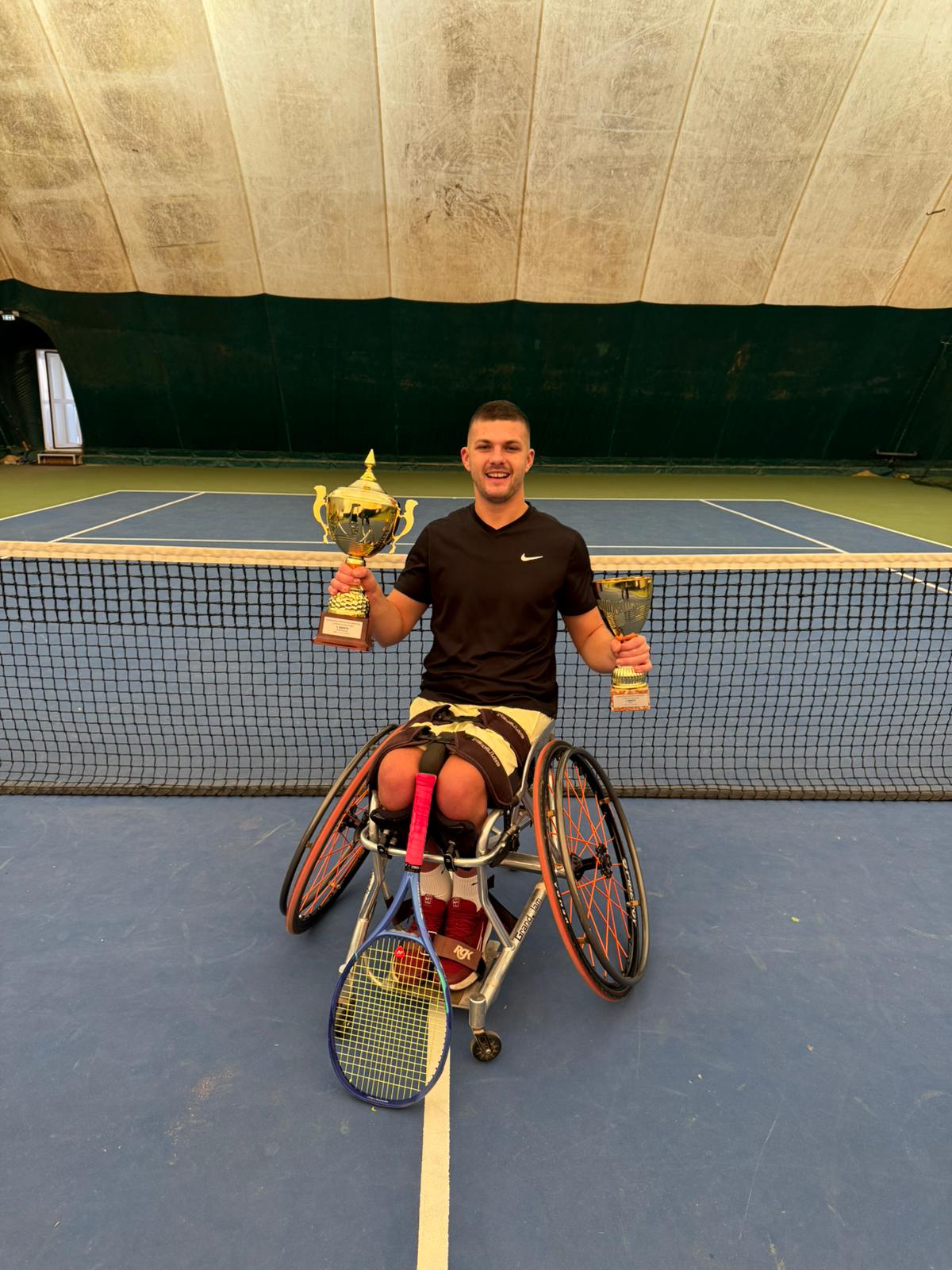 Slika prikazuje tenisača u kolicima kako drži dva pehara, a reket mu je naslonjen na noge. On se nalazi na teniskom terenu, a iza njega je vidljiva teniska mreža.