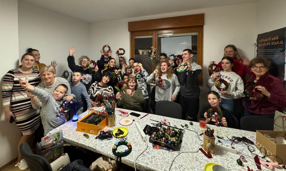 Na slici se nalazi vesela skupina ljudi okupljena u zatvorenom prostoru, sudjelujući u blagdanskoj radionici izrade božićnih ukrasa. Sudionici, među kojima su djeca, odrasli i stariji, ponosno drže ručno izrađene adventske vijence i dekoracije, pozirajući s osmijehom za zajedničku fotografiju. Na stolu ispred njih nalaze se razni materijali za kreativni rad: škare, vrpce, kuglice, pištolji za ljepilo i ukrasni elementi. Atmosfera je topla i zajednička, ispunjena blagdanskim duhom, kreativnošću i suradnjom. Na zidu u pozadini vidi se plakat s tekstom na slavenskom jeziku, najvjerojatnije hrvatskom ili srpskom, što daje dodatni kulturni kontekst događaju. Sve skupa odaje dojam inkluzivne i razigrane radionice, vjerojatno organizirane u lokalnoj zajednici povodom božićnih blagdana.