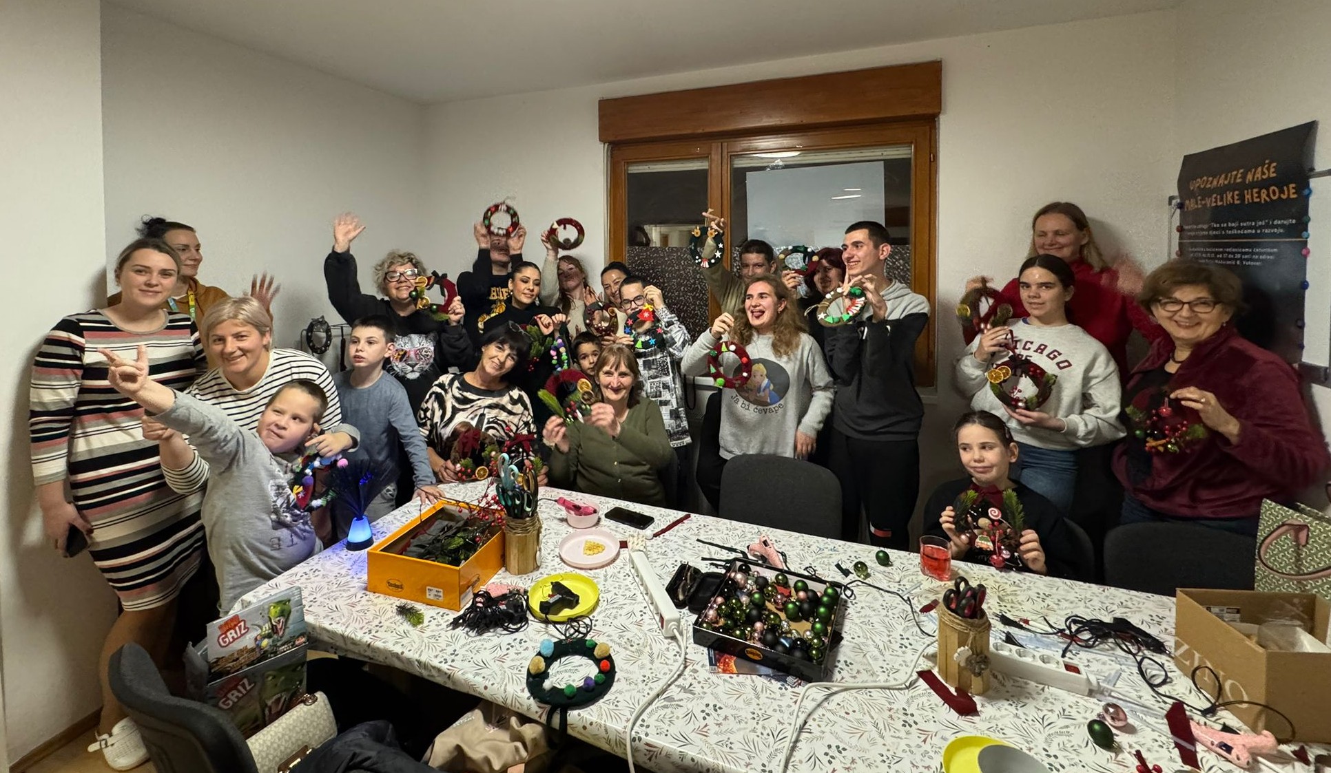 Na slici se nalazi vesela skupina ljudi okupljena u zatvorenom prostoru, sudjelujući u blagdanskoj radionici izrade božićnih ukrasa. Sudionici, među kojima su djeca, odrasli i stariji, ponosno drže ručno izrađene adventske vijence i dekoracije, pozirajući s osmijehom za zajedničku fotografiju. Na stolu ispred njih nalaze se razni materijali za kreativni rad: škare, vrpce, kuglice, pištolji za ljepilo i ukrasni elementi. Atmosfera je topla i zajednička, ispunjena blagdanskim duhom, kreativnošću i suradnjom. Na zidu u pozadini vidi se plakat s tekstom na slavenskom jeziku, najvjerojatnije hrvatskom ili srpskom, što daje dodatni kulturni kontekst događaju. Sve skupa odaje dojam inkluzivne i razigrane radionice, vjerojatno organizirane u lokalnoj zajednici povodom božićnih blagdana.