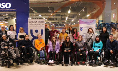 Velika skupina članova i volontera Udruge osoba s mišićnom distrofijom Primorsko-goranske županije pozira u trgovačkom centru ispred dva promotivna roll-up plakata udruge. U prvom redu sjede osobe u invalidskim kolicima, dok iza njih stoje članovi udruge i volonteri, neki drže poklon-paketiće. Okruženje je svijetlo, s vidljivim oznakama trgovina u pozadini. Atmosfera je topla, zajednička i svečana u povodu obilježavanja Međunarodnog dana osoba s invaliditetom.