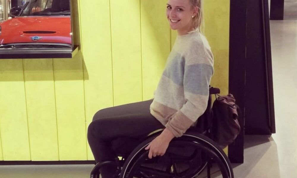 Mlada žena u invalidskim kolicima nasmiješeno pozira u unutarnjem prostoru. Odjevena je u svjetli prugasti pulover i tamne hlače, a iza nje je zid obložen žutim drvenim panelima. Uz rub kadra vidi se dio izloga s crvenim automobilom. Atmosfera je opuštena i svakodnevna.