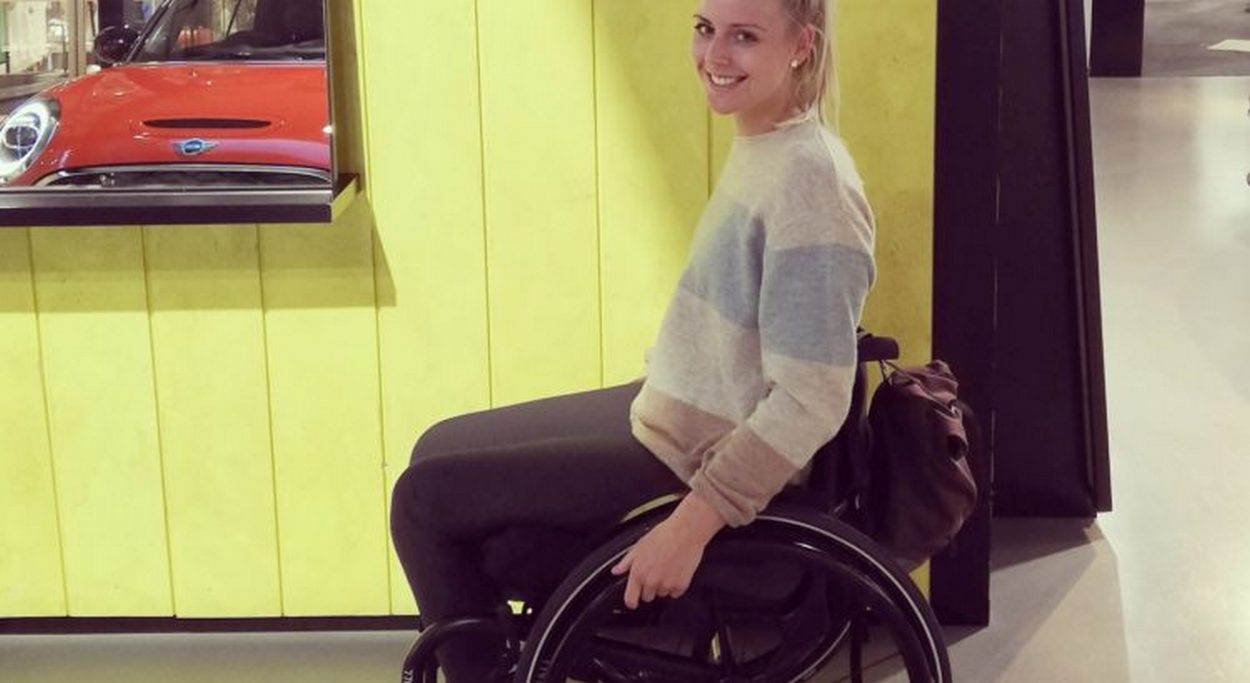 Mlada žena u invalidskim kolicima nasmiješeno pozira u unutarnjem prostoru. Odjevena je u svjetli prugasti pulover i tamne hlače, a iza nje je zid obložen žutim drvenim panelima. Uz rub kadra vidi se dio izloga s crvenim automobilom. Atmosfera je opuštena i svakodnevna.