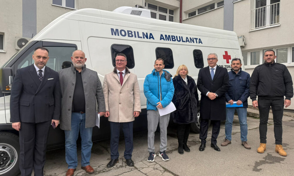 Na fotografiji se nalazi osam osoba koje stoje ispred bijelog mobilnog medicinskog vozila s natpisom „MOBILNA AMBULANTA”, što znači „mobilna ambulanta”. Na vozilu je vidljiv i crveni križ te tekst „DOM ZDRAVLJA”, koji označava da je vozilo povezano sa zdravstvenom ustanovom.