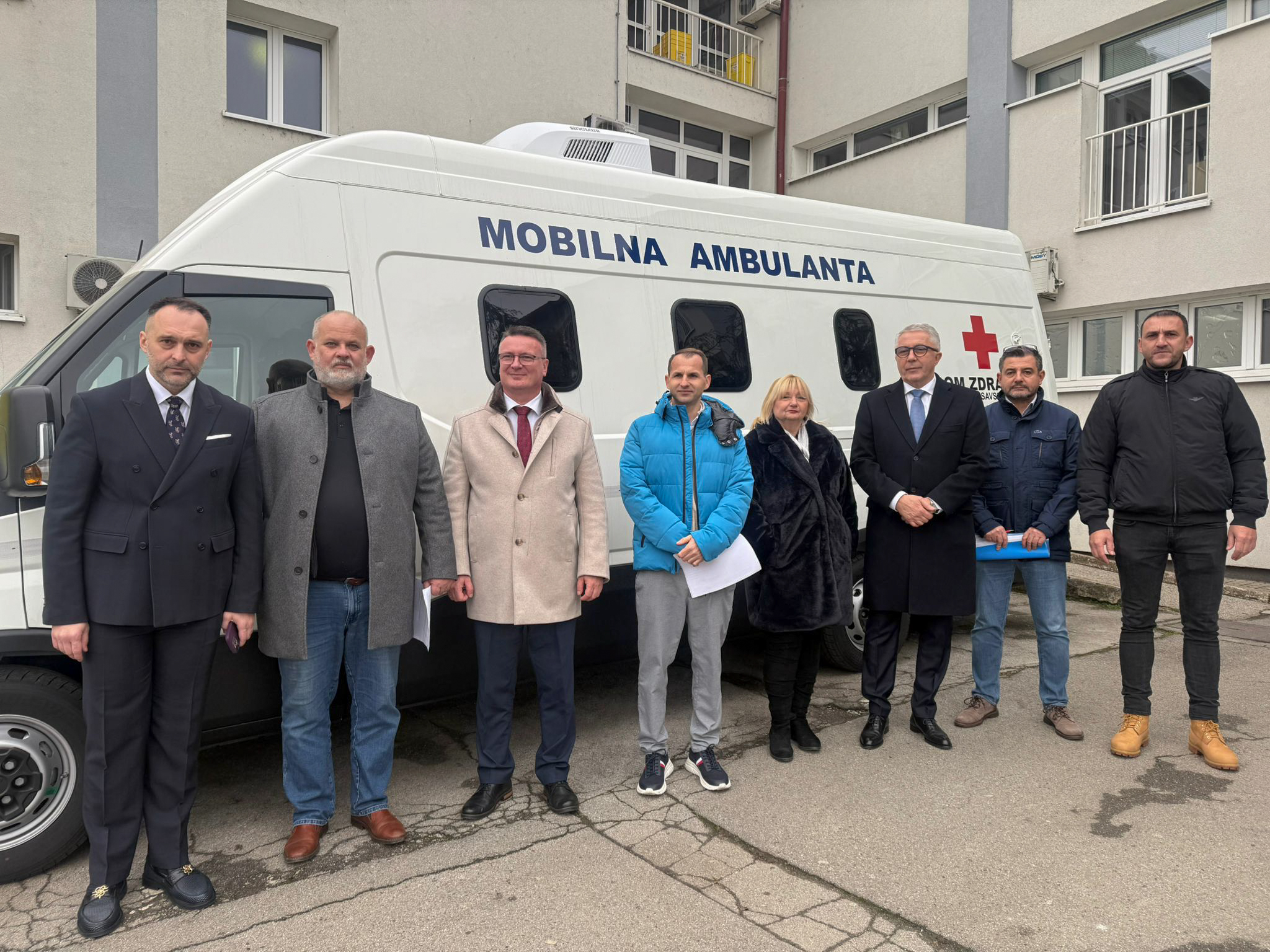 Na fotografiji se nalazi osam osoba koje stoje ispred bijelog mobilnog medicinskog vozila s natpisom „MOBILNA AMBULANTA”, što znači „mobilna ambulanta”. Na vozilu je vidljiv i crveni križ te tekst „DOM ZDRAVLJA”, koji označava da je vozilo povezano sa zdravstvenom ustanovom.