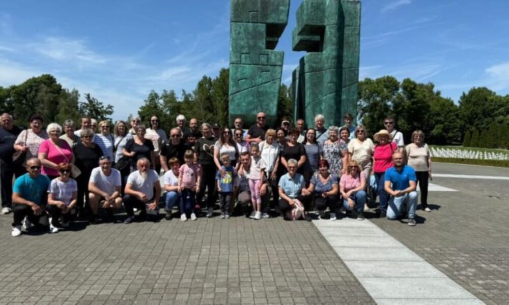 Na fotografiji je prikazana velika skupina ljudi različitih generacija – od djece do starijih osoba – okupljena ispred monumentalne skulpture. Skulptura se sastoji od dvaju visokih, apstraktnih brončanih elemenata zelenkaste patine, s geometrijskim oblicima i otvorima, postavljenih na popločanoj površini. Njezin oblik i smještaj sugeriraju da se radi o spomeniku s povijesnim ili memorijalnim značenjem. U pozadini se vidi vedro plavo nebo, krošnje drveća i polje bijelih oznaka ili nadgrobnih ploča, što dodatno potvrđuje da je lokacija vjerojatno memorijalno mjesto. Atmosfera fotografije odiše dostojanstvom, zajedništvom i kolektivnim prisjećanjem – trenutak okupljanja koji nosi emocionalnu težinu i kulturnu važnost.