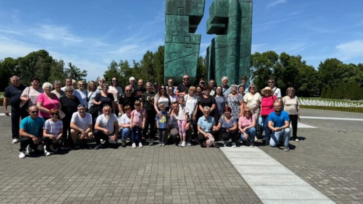 Na fotografiji je prikazana velika skupina ljudi različitih generacija – od djece do starijih osoba – okupljena ispred monumentalne skulpture. Skulptura se sastoji od dvaju visokih, apstraktnih brončanih elemenata zelenkaste patine, s geometrijskim oblicima i otvorima, postavljenih na popločanoj površini. Njezin oblik i smještaj sugeriraju da se radi o spomeniku s povijesnim ili memorijalnim značenjem. U pozadini se vidi vedro plavo nebo, krošnje drveća i polje bijelih oznaka ili nadgrobnih ploča, što dodatno potvrđuje da je lokacija vjerojatno memorijalno mjesto. Atmosfera fotografije odiše dostojanstvom, zajedništvom i kolektivnim prisjećanjem – trenutak okupljanja koji nosi emocionalnu težinu i kulturnu važnost.