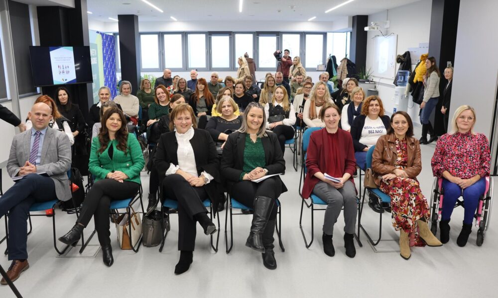 Na fotografiji je prikazana skupina ljudi koji sjede u modernom, svijetlom prostoru – najvjerojatnije sudjeluju na seminaru, radionici ili sastanku. Prostorija ima velika prozorska stakla koja propuštaju puno prirodnog svjetla, a sudionici su raspoređeni u redovima, okrenuti prema kameri. Na lijevoj strani vidljiv je ekran s prezentacijom na kojem piše „What do we do in our free time?“ uz ilustrirane figure, dok je projektor montiran na stropu. Sudionici djeluju angažirano i pripremljeno – neki drže bilježnice ili papire. U prvom redu sjede osobe koje bi mogle biti govornici ili organizatori, s obzirom na njihov položaj i odjeću. Jedna osoba u drugom redu nosi majicu s natpisom „NIŠTA O NAMA BEZ NAS!“, što upućuje na temu inkluzije i participacije.