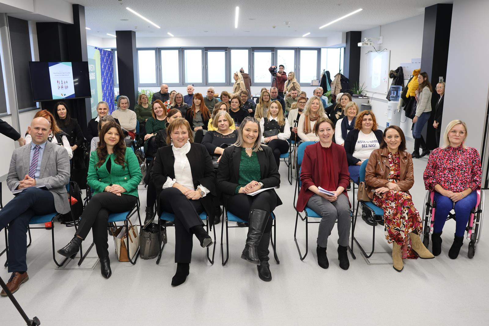 Na fotografiji je prikazana skupina ljudi koji sjede u modernom, svijetlom prostoru – najvjerojatnije sudjeluju na seminaru, radionici ili sastanku. Prostorija ima velika prozorska stakla koja propuštaju puno prirodnog svjetla, a sudionici su raspoređeni u redovima, okrenuti prema kameri. Na lijevoj strani vidljiv je ekran s prezentacijom na kojem piše „What do we do in our free time?“ uz ilustrirane figure, dok je projektor montiran na stropu. Sudionici djeluju angažirano i pripremljeno – neki drže bilježnice ili papire. U prvom redu sjede osobe koje bi mogle biti govornici ili organizatori, s obzirom na njihov položaj i odjeću. Jedna osoba u drugom redu nosi majicu s natpisom „NIŠTA O NAMA BEZ NAS!“, što upućuje na temu inkluzije i participacije.