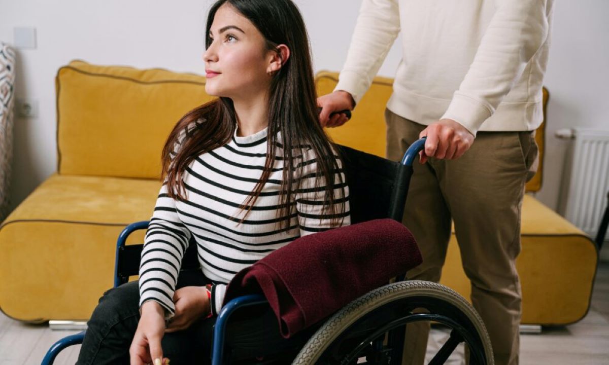 Na slici se nalazi osoba u invalidskim kolicima, odjevena u crno-bijelu prugastu majicu dugih rukava, s dugom tamnom kosom. Preko naslona kolica prebačena je bordo deka, koja unosi toplinu u prizor. Iza nje stoji druga osoba, obučena u bijeli pulover i smeđe hlače, koja drži ručke kolica – njezin stav izražava pažnju i spremnost na pomoć. Okruženje je intimno i domaće: u pozadini se vidi senf-žuti kauč i radijator, što sugerira unutarnji prostor, vjerojatno dnevni boravak. Svjetlo je blago, a atmosfera mirna i podržavajuća.