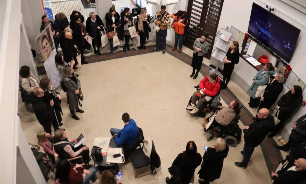 Fotografija prikazuje skupinu ljudi okupljenih u kružnoj formaciji unutar prostrane, svijetle zgrade — moguće društvenog centra, galerije ili edukativnog prostora. Sudionici sjede i stoje, raspoređeni oko središnjeg dijela, gdje se odvija prezentacija ili razgovor. Među prisutnima su i dvije osobe u invalidskim kolicima, što jasno naglašava inkluzivnost i pristupačnost događaja. Prostor je uredan, s pločicama na podu, velikim ekranom na zidu i informativnim panoima. Mnogi sudionici drže platnene torbe, a neki su zaokupljeni mobilnim uređajima ili tiskanim materijalima. Na lijevoj strani fotografije vidljiv je banner s portretom žene i tekstom, što upućuje na promotivni ili edukativni sadržaj. Opći dojam fotografije je toplina zajedništva, otvorenost i aktivno sudjelovanje. Prikazuje trenutak u kojem se različiti članovi zajednice okupljaju radi razmjene znanja, podrške ili informiranja — s jasnim naglaskom na uključivanje osoba s invaliditetom.