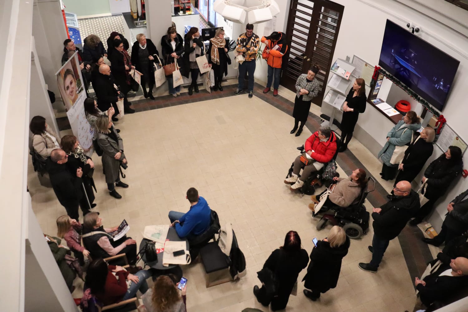 Fotografija prikazuje skupinu ljudi okupljenih u kružnoj formaciji unutar prostrane, svijetle zgrade — moguće društvenog centra, galerije ili edukativnog prostora. Sudionici sjede i stoje, raspoređeni oko središnjeg dijela, gdje se odvija prezentacija ili razgovor. Među prisutnima su i dvije osobe u invalidskim kolicima, što jasno naglašava inkluzivnost i pristupačnost događaja. Prostor je uredan, s pločicama na podu, velikim ekranom na zidu i informativnim panoima. Mnogi sudionici drže platnene torbe, a neki su zaokupljeni mobilnim uređajima ili tiskanim materijalima. Na lijevoj strani fotografije vidljiv je banner s portretom žene i tekstom, što upućuje na promotivni ili edukativni sadržaj. Opći dojam fotografije je toplina zajedništva, otvorenost i aktivno sudjelovanje. Prikazuje trenutak u kojem se različiti članovi zajednice okupljaju radi razmjene znanja, podrške ili informiranja — s jasnim naglaskom na uključivanje osoba s invaliditetom.