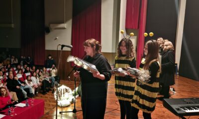 Fotografija prikazuje svečani trenutak na pozornici u kazališnoj ili školskoj dvorani. U prvom planu stoji žena s mikrofonom, držeći buket cvijeća omotan u ukrasni papir — vjerojatno u znak zahvale ili priznanja. Pokraj nje stoje dvoje djece odjevene u kostime pčela, također s buketima u rukama, što sugerira da su sudjelovali u nekoj izvedbi ili programu.