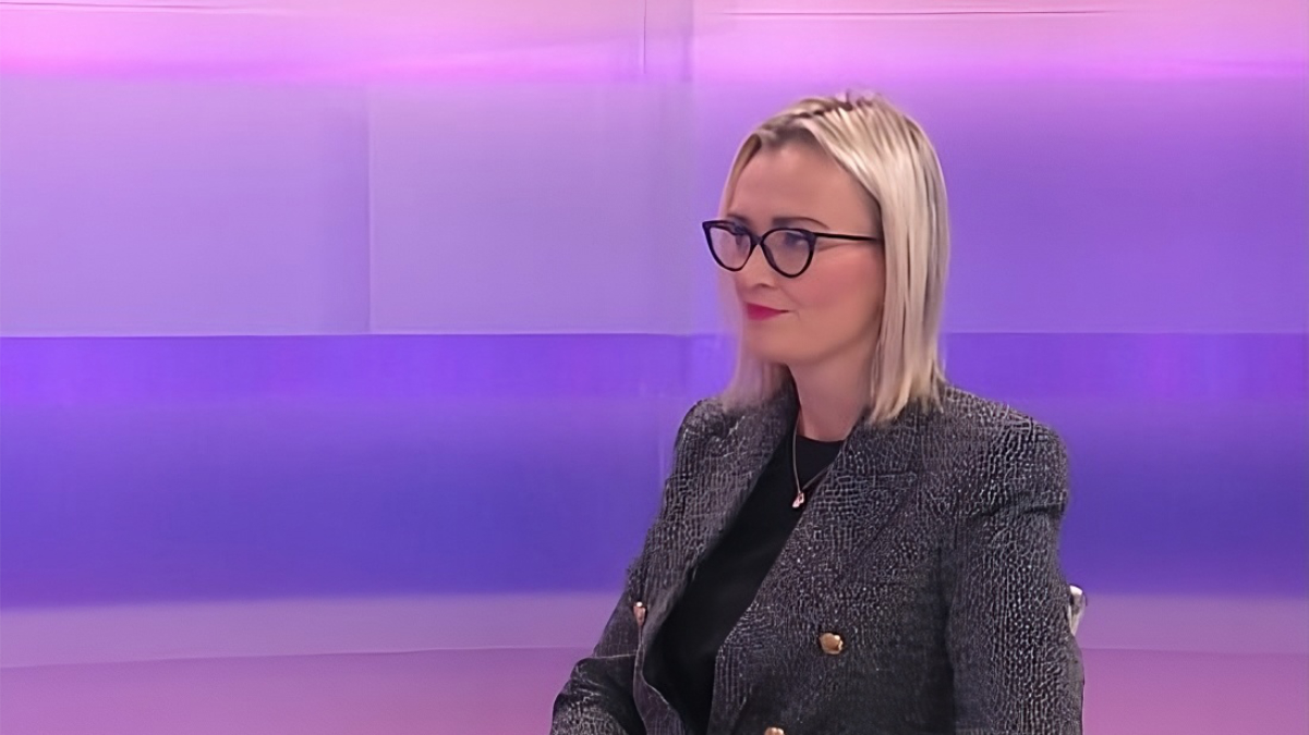 Žena svijetle kose i naočala sjedi u televizijskom studiju s ljubičasto osvijetljenom pozadinom. Odjevena je u tamni sako s dvorednim kopčanjem i crnu majicu, gleda blago u stranu i djeluje smireno i fokusirano, kao tijekom intervjua ili studijskog razgovora.