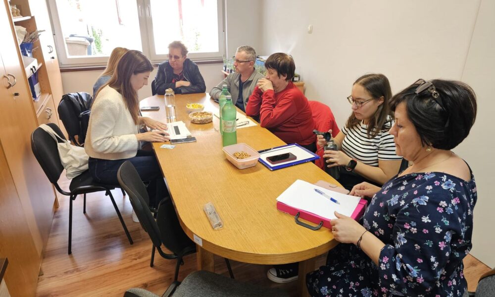 Na fotografiji je prikazana skupina od sedam osoba okupljenih oko drvenog konferencijskog stola u manjoj prostoriji za sastanke. Prostor ima drveni pod, bijele zidove i veliki prozor kroz koji dopire prirodna svjetlost, stvarajući ugodnu i svijetlu atmosferu. Na stolu se nalaze razni predmeti: bilježnice, olovke, laptop, boca vode, zelena boca s oznakom „Jamnica“, zdjelice s grickalicama i ravnalo. Neki sudionici razgovaraju, dok drugi pišu bilješke ili tipkaju na laptopu. Cjelokupna scena sugerira da se radi o suradničkom sastanku – možda planiranju projekta, edukaciji ili organizacijskom dogovoru.