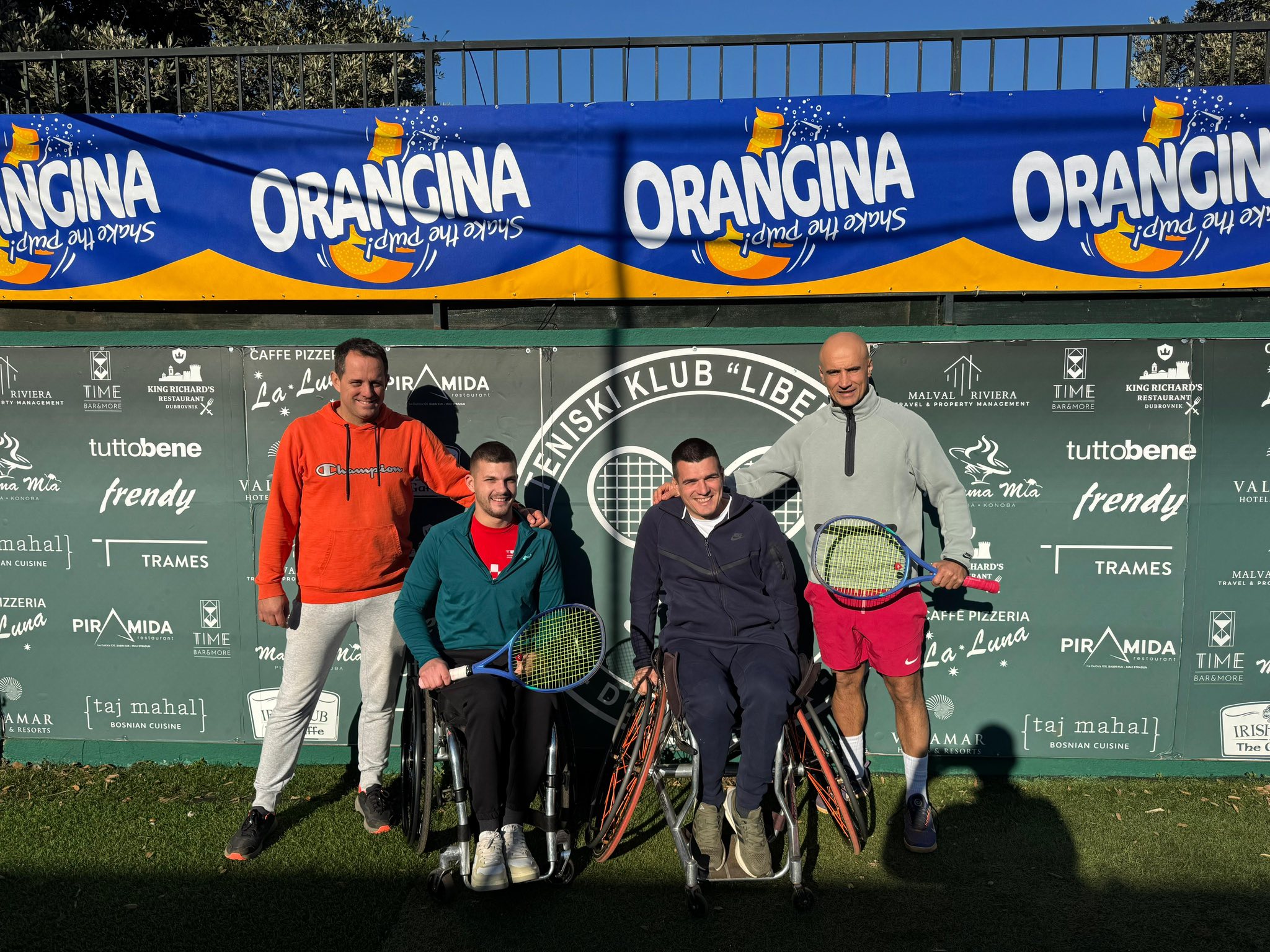 Fotografija prikazuje tenisače u kolicima i jednog čovjeka koji stoji iza njih.