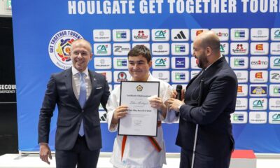 Na fotografiji je prikazana svečana dodjela nagrade tijekom Europske judo turneje “Houlgate Get Together”. U prvom planu stoje tri osobe: Mladi judo sportaš u bijelom kimonu s narančasto-zelenim pojasom, ponosno drži certifikat o postignuću. S njegove lijeve i desne strane nalaze se dva muškarca u svečanoj odjeći, od kojih jedan drži mikrofon – vjerojatno voditelj ceremonije ili predstavnik organizacije. U pozadini se nalazi plavi banner s nazivom događaja i logotipima sponzora, uključujući CA Normandie, Adidas, Mizuno i druge. Vizualna kompozicija jasno ističe trenutak priznanja, dok boje i raspored elemenata stvaraju profesionalan i svečan dojam. 📜 Tekst na certifikatu koji drži sportaš glasi: Certificate of Achievement Tiber Andgjar EJU Fair Play Award of 2024 Potpis: Dr. T. Toth, EJU Vice President Datum: prosinac 2024.