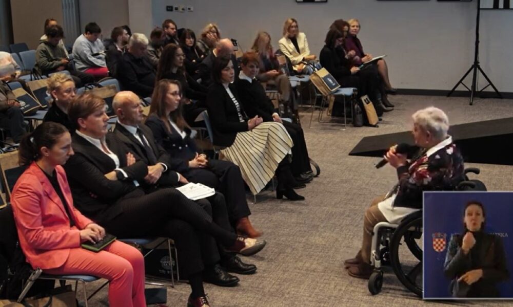 a fotografiji je prikazana konferencijska ili seminarska dvorana ispunjena publikom koja pažljivo sluša govornika. Govornik je starija osoba u invalidskim kolicima, drži mikrofon i obraća se prisutnima. Publika je raznolika – ljudi različitih dobnih skupina, odjeveni u formalnu i poluformalnu odjeću, sjede u redovima stolica. Na mnogim stolicama vise platnene torbe, vjerojatno s materijalima za konferenciju. U donjem desnom kutu fotografije nalazi se umetnuta slika tumača za znakovni jezik, ispred plave pozadine s hrvatskim grbom i natpisom „REPUBLIKA HRVATSKA“, što upućuje na to da se događaj odvija u Hrvatskoj ili je povezan s hrvatskim institucijama. Fotografija snažno ističe inkluzivnost i pristupačnost javnih događanja – prisutnost govornika u invalidskim kolicima te osiguranje tumačenja na znakovni jezik za osobe s oštećenjem sluha.
