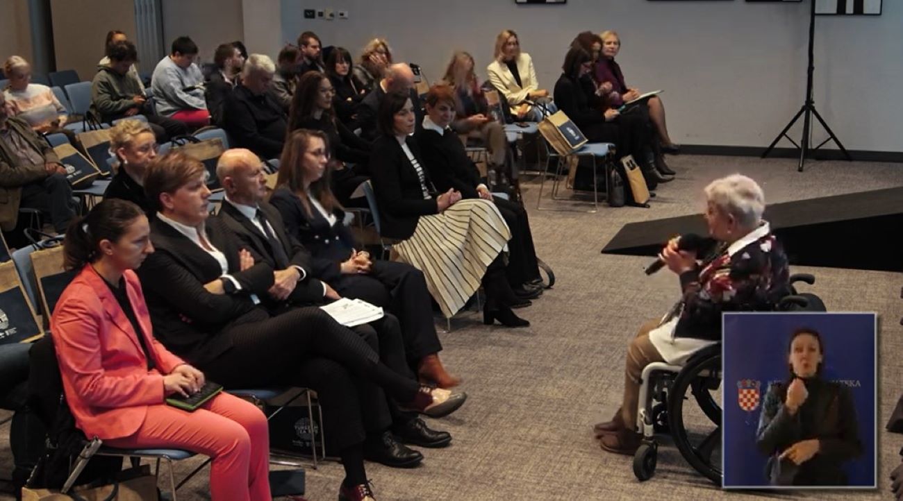 a fotografiji je prikazana konferencijska ili seminarska dvorana ispunjena publikom koja pažljivo sluša govornika. Govornik je starija osoba u invalidskim kolicima, drži mikrofon i obraća se prisutnima. Publika je raznolika – ljudi različitih dobnih skupina, odjeveni u formalnu i poluformalnu odjeću, sjede u redovima stolica. Na mnogim stolicama vise platnene torbe, vjerojatno s materijalima za konferenciju. U donjem desnom kutu fotografije nalazi se umetnuta slika tumača za znakovni jezik, ispred plave pozadine s hrvatskim grbom i natpisom „REPUBLIKA HRVATSKA“, što upućuje na to da se događaj odvija u Hrvatskoj ili je povezan s hrvatskim institucijama. Fotografija snažno ističe inkluzivnost i pristupačnost javnih događanja – prisutnost govornika u invalidskim kolicima te osiguranje tumačenja na znakovni jezik za osobe s oštećenjem sluha.