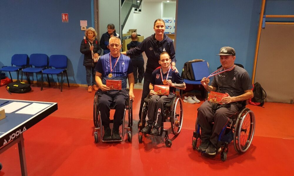 Fotografija prikazuje troje ljudi u invalidskim kolicima i ženu koja stoji iza njih. To troje ljudi u kolicima imaju medalje oko vrata. Nalaze se u nekoj dvorani, a u pozadini se vidi dio stola za stolni tenis.