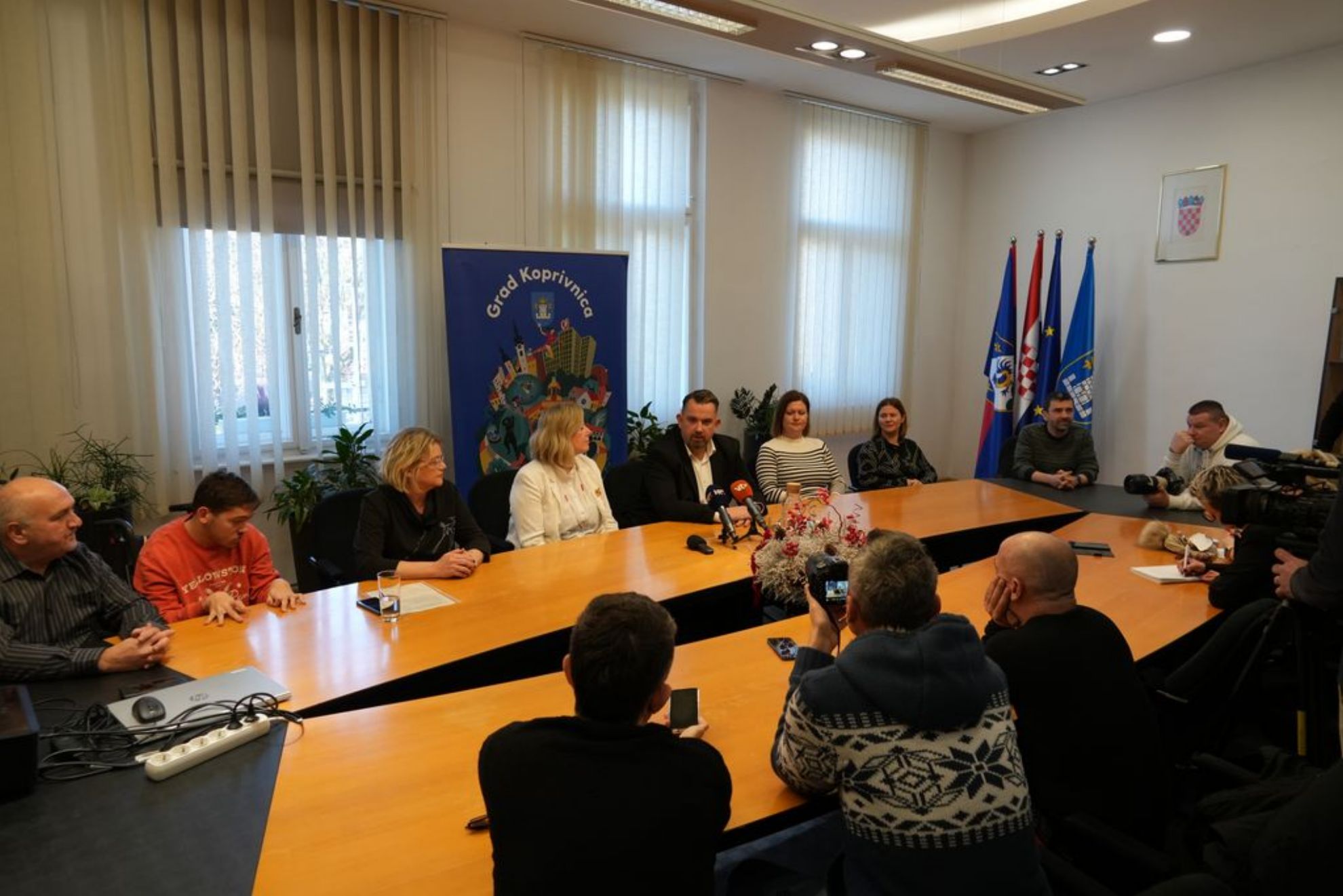 Fotografija prikazuje službeni sastanak ili konferenciju za medije u svijetloj prostoriji s velikim prozorima i vertikalnim žaluzinama. Sudionici su okupljeni oko velikog drvenog konferencijskog stola, na kojem se nalaze mikrofoni i uređaji za snimanje, što upućuje na to da se događaj dokumentira ili prenosi. Na jednoj strani stola sjede osobe koje izgledaju kao dužnosnici ili govornici, dok su s druge strane prisutni novinari i fotografi, neki sjede, a neki stoje i snimaju događaj. U pozadini se nalazi plavi banner s natpisom „Grad Koprivnica” i ilustracijom grada, što sugerira da se događaj odvija u Koprivnici, Hrvatska. Iza bannera stoje tri zastave: hrvatska nacionalna zastava, gradska ili županijska zastava te zastava Europske unije. Na zidu je i uokvireni grb Republike Hrvatske. Cjelokupna atmosfera fotografije odaje dojam važnog javnog događaja, moguće vezanog uz lokalnu upravu, zajedničke inicijative ili objave za javnost.