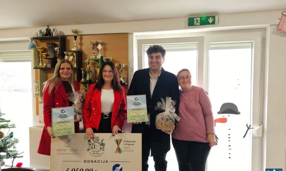 Fotografija prikazuje svečanu prigodu u zatvorenom prostoru, vjerojatno u sklopu humanitarnog ili božićnog događanja. Četiri odrasle osobe stoje ispred zida s trofejima i prozorom ukrašenim snjegovićem. Svi su nasmijani i drže zahvalnice, darovne košare te veliki simbolični ček na iznos od 5.050,00 eura. Na čeku piše da je donacija namijenjena Udruzi „Maslačak“ iz Križevaca, a prisutni su predstavnici organizacija — dvije žene u crvenim sakooma, muškarac u crnom kaputu i žena u ružičastoj majici. U pozadini se vidi božićno drvce, što dodatno naglašava blagdanski ugođaj i duh darivanja. Cijela scena odiše toplinom, solidarnošću i zajedništvom, a fotografija bilježi trenutak zahvale i podrške lokalnoj zajednici.