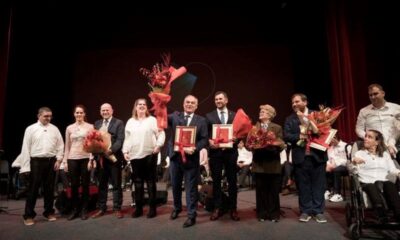 Na fotografiji je prikazana svečana dodjela priznanja koja se odvija na pozornici s tamnom pozadinom i crvenim zavjesama sa strane. U prvom planu stoji skupina ljudi odjevenih u svečanu i polusvečanu odjeću. Nekoliko osoba drži velike bukete cvijeća i uokvirene nagrade, što jasno upućuje na trenutak priznanja i slavljenja postignuća. 🎖️ Među nagrađenima i sudionicima vidljive su osobe s invaliditetom, što snažno naglašava inkluzivnost i poštovanje prema različitostima. U pozadini se nalaze dodatni sudionici ili publika, smješteni u sjedećem položaju, što dodatno potvrđuje formalni karakter događaja. Cjelokupna atmosfera odiše toplinom, dostojanstvom i zajedništvom, a fotografija bilježi važan trenutak priznanja doprinosa u području koje vjerojatno uključuje zagovaranje prava osoba s invaliditetom, društvenu odgovornost ili kulturnu raznolikost.