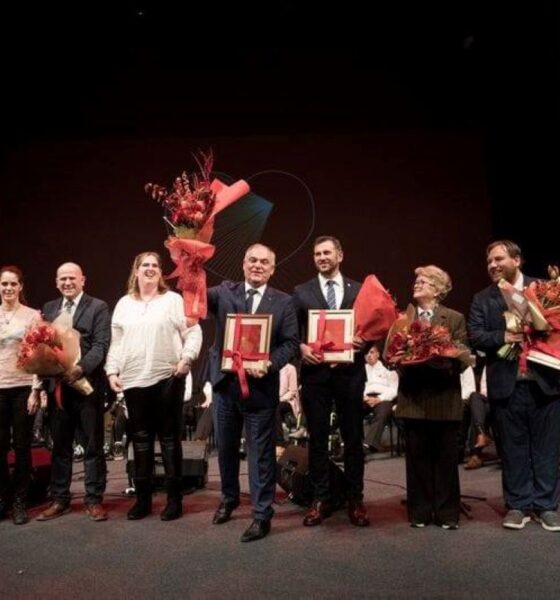 Na fotografiji je prikazana svečana dodjela priznanja koja se odvija na pozornici s tamnom pozadinom i crvenim zavjesama sa strane. U prvom planu stoji skupina ljudi odjevenih u svečanu i polusvečanu odjeću. Nekoliko osoba drži velike bukete cvijeća i uokvirene nagrade, što jasno upućuje na trenutak priznanja i slavljenja postignuća. 🎖️ Među nagrađenima i sudionicima vidljive su osobe s invaliditetom, što snažno naglašava inkluzivnost i poštovanje prema različitostima. U pozadini se nalaze dodatni sudionici ili publika, smješteni u sjedećem položaju, što dodatno potvrđuje formalni karakter događaja. Cjelokupna atmosfera odiše toplinom, dostojanstvom i zajedništvom, a fotografija bilježi važan trenutak priznanja doprinosa u području koje vjerojatno uključuje zagovaranje prava osoba s invaliditetom, društvenu odgovornost ili kulturnu raznolikost.