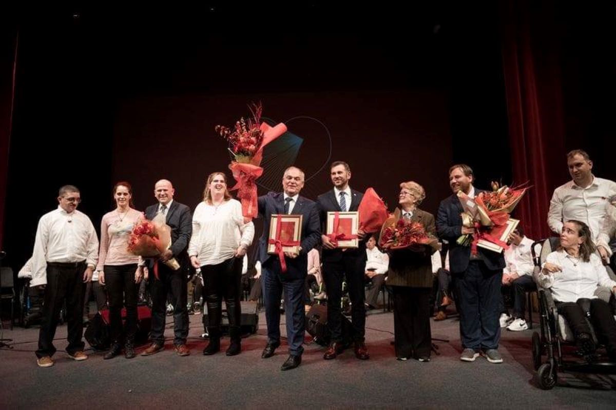 Na fotografiji je prikazana svečana dodjela priznanja koja se odvija na pozornici s tamnom pozadinom i crvenim zavjesama sa strane. U prvom planu stoji skupina ljudi odjevenih u svečanu i polusvečanu odjeću. Nekoliko osoba drži velike bukete cvijeća i uokvirene nagrade, što jasno upućuje na trenutak priznanja i slavljenja postignuća. 🎖️ Među nagrađenima i sudionicima vidljive su osobe s invaliditetom, što snažno naglašava inkluzivnost i poštovanje prema različitostima. U pozadini se nalaze dodatni sudionici ili publika, smješteni u sjedećem položaju, što dodatno potvrđuje formalni karakter događaja. Cjelokupna atmosfera odiše toplinom, dostojanstvom i zajedništvom, a fotografija bilježi važan trenutak priznanja doprinosa u području koje vjerojatno uključuje zagovaranje prava osoba s invaliditetom, društvenu odgovornost ili kulturnu raznolikost.