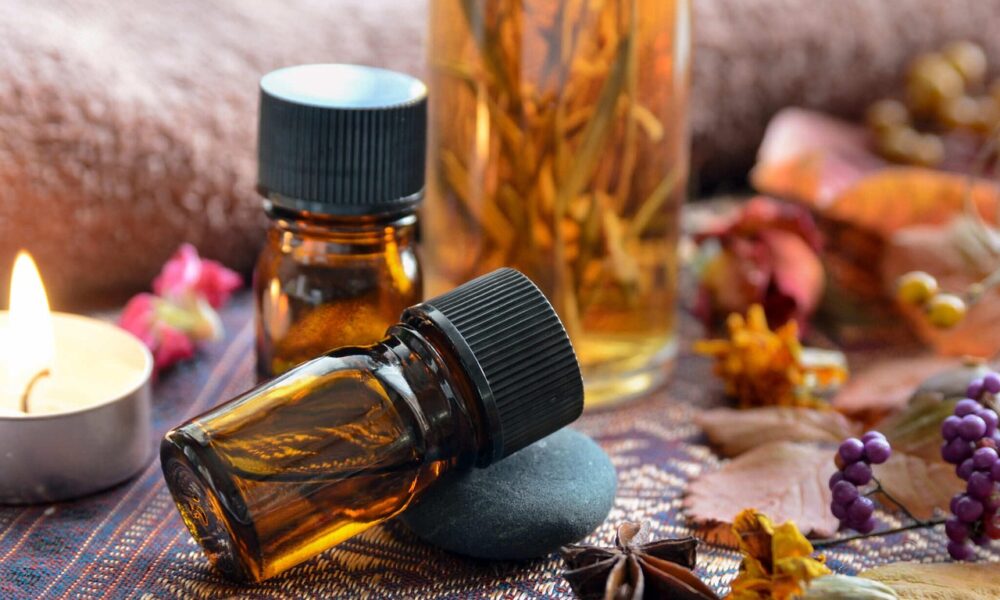 Na fotografiji je prikazana smirujuća i estetski skladna scena s jantarnim staklenim bočicama, vjerojatno s eteričnim uljima, koje su postavljene na tkaninu s uzorkom. Jedna bočica leži na glatkom crnom kamenu, dok druga stoji uspravno. U pozadini se nalazi veća bočica s biljem ili biljnim materijalom uronjenim u tekućinu, što sugerira domaću infuziju ili dekorativno ulje. Oko bočica su raspoređeni prirodni elementi poput osušenog cvijeća, zvjezdastog anisa, ljubičastih bobica i jesenskog lišća, koji pridonose toploj i zemljanoj atmosferi. Upaljena lučica dodaje nježan sjaj, pojačavajući dojam mira i ambijenta sličnog spa centru. Ova slika savršeno dočarava teme aromaterapije, opuštanja, prirodnih pripravaka i wellnessa.