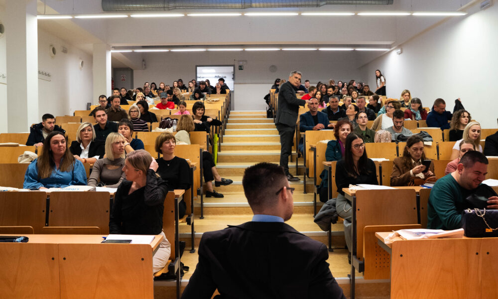 Na fotografiji je prikazana velika predavaonica ispunjena ljudima koji sjede u redovima drvenih klupa. Prostorija je svijetla, s bijelim zidovima i stropom, a središnje stubište dijeli sjedeći dio na dvije strane. U prvom planu jasno se vidi osoba u invalidskim kolicima, okrenuta prema prednjem dijelu dvorane. Na sredini stubišta stoji muškarac koji izgleda kao da se obraća publici, što sugerira da je u tijeku predavanje ili prezentacija. Publika je raznolika i djeluje pažljivo, neki prate izlaganje, dok drugi rade s materijalima ispred sebe.