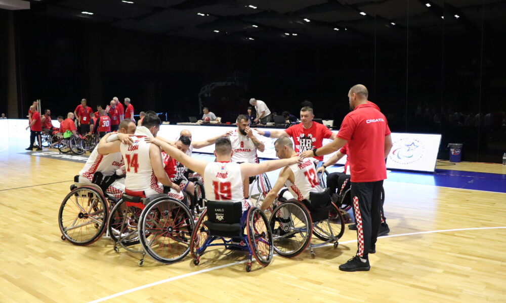 Slika prikazuje nekoliko igrača u košarkaškim dresovima kako sjede u invalidskim kolicima poredani u krug.