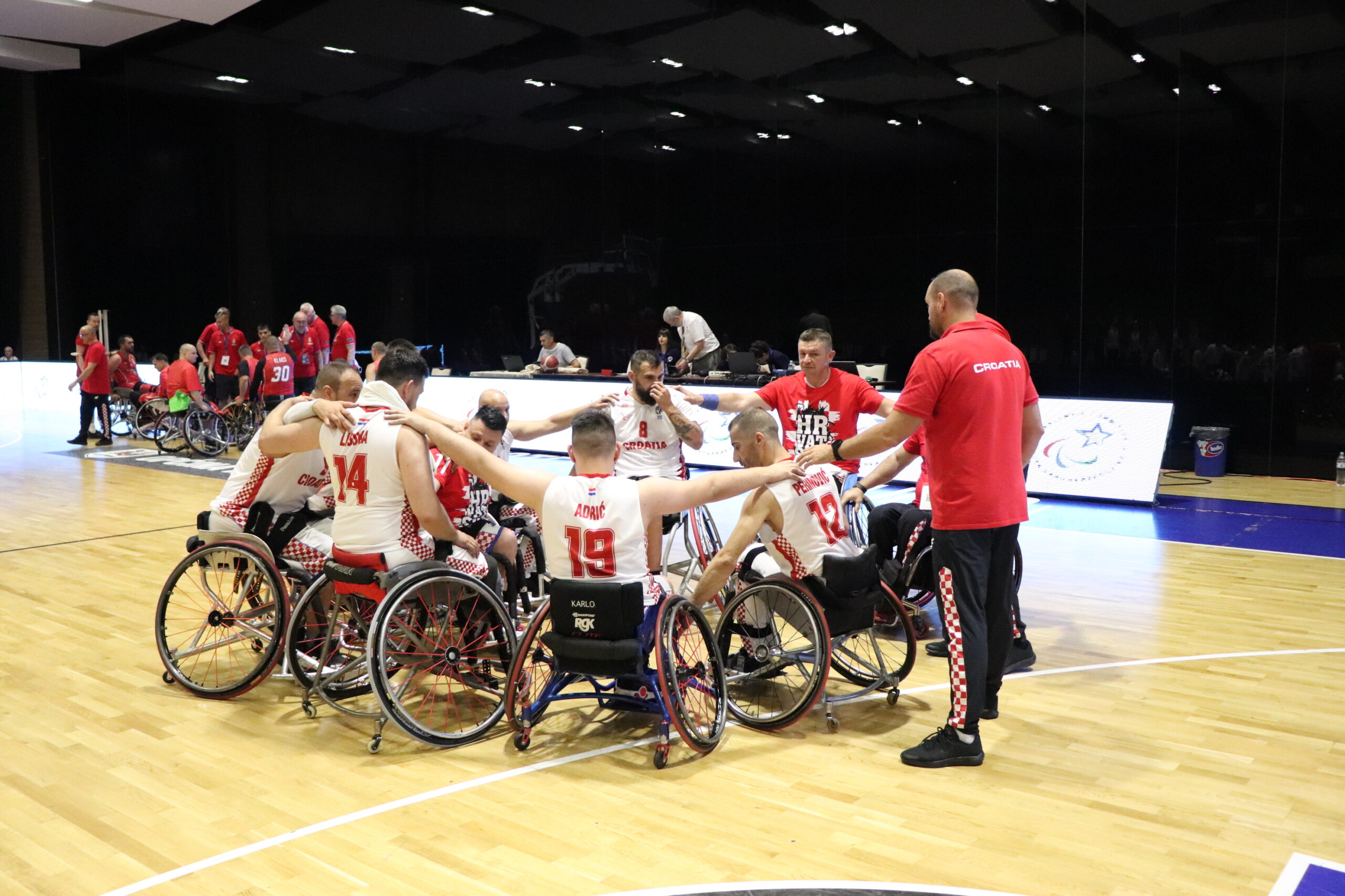 Slika prikazuje nekoliko igrača u košarkaškim dresovima kako sjede u invalidskim kolicima poredani u krug.