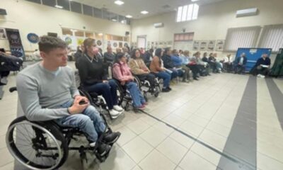 Na fotografiji se nalazi skupina ljudi okupljena u velikoj zatvorenoj prostoriji koja izgleda kao društveni centar ili dvorana za sastanke. Nekoliko osoba u prvom redu sjedi u invalidskim kolicima, što sugerira da je događaj povezan s pristupačnošću, podizanjem svijesti o invaliditetu ili podrškom osobama s poteškoćama u kretanju. Prostorija ima pločice na podu, fluorescentnu rasvjetu i zidove ukrašene uokvirenim slikama ili plakatima. Publika je usmjerena prema prednjem dijelu prostorije, gdje su dvije osobe smještene i vjerojatno vode raspravu ili prezentaciju. Atmosfera djeluje ozbiljno i angažirano, a fotografija ističe važnost inkluzije i zajedničkog djelovanja u stvaranju pristupačnog okruženja za sve članove društva.