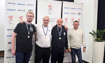 Četvorica muškaraca stoje ispred zida s logotipima Hrvatskog paraolimpijskog odbora i sponzora. Trojica u sredini nose medalje – brončanu, zlatnu i srebrnu – i nasmiješeno stoje jedan uz drugoga s rukama preko ramena. Četvrti muškarac, u bijelom džemperu, stoji desno bez medalje. Atmosfera je svečana i sportska.