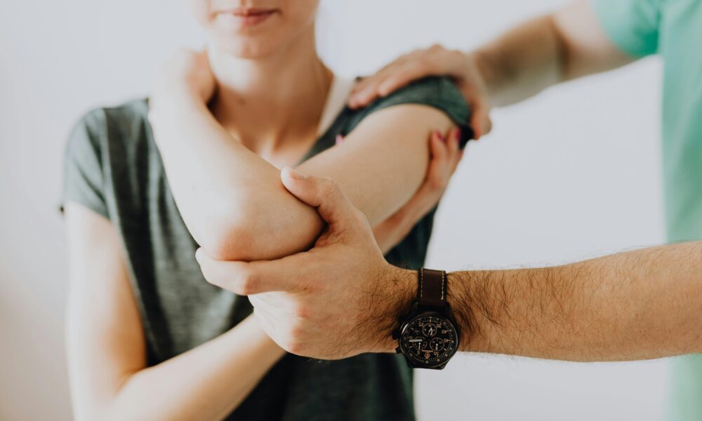 Fotografija prikazuje trenutak iz fizikalne terapije ili rehabilitacijskog tretmana. U prvom planu vidimo osobu koja pruža stručnu pomoć drugoj osobi — jednom rukom podupire njezin lakat, a drugom rame, što upućuje na vježbu mobilnosti ili istezanja. Osoba koja prima pomoć nosi tamnosivu majicu, dok terapeut ili asistent ima zelenu majicu i sat na ruci. Pokret je pažljiv i usmjeren na podršku, što sugerira profesionalan i empatičan pristup u radu s pacijentima.