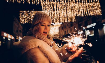 Fotografija prikazuje osobu odjevenu u toplu zimsku odjeću: pletenu kapu, šal i kaput s krznenim rubom. U ruci drži upaljenu prskalicu koja svijetli u tamnijem ambijentu, stvarajući iskričav, svečan ugođaj. Pozadina je ispunjena visećim svjetlosnim dekoracijama koje dodatno naglašavaju blagdansku atmosferu — vjerojatno u urbanom okruženju tijekom večernjih sati. Toplina svjetla prskalice i ambijentalnih lampica stvara osjećaj radosti, zajedništva i zimskog slavlja. Fotografija odiše blagdanskim duhom, a može se povezati s proslavama Božića, Nove godine ili zimskih festivala. Osoba na slici djeluje zaigrano i prisutno, uhvaćena u trenutku osobne radosti.