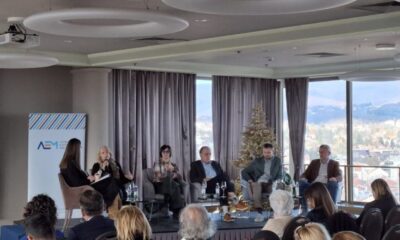 Na slici je prikazana profesionalna panel rasprava ili konferencija koja se odvija u modernom zatvorenom prostoru s velikim staklenim prozorima. Kroz njih se pruža panoramski pogled na grad i okolna brda, što prostoru daje svijetlu i otvorenu atmosferu. Na pozornici sjedi šest osoba u polukrugu, uključujući jednu osobu koja aktivno govori u mikrofon. Svi sudionici djeluju koncentrirano i angažirano, što sugerira ozbiljnu i sadržajnu diskusiju. Publika je smještena u redovima ispred pozornice, pažljivo prateći događaj. Na pozornici se nalazi i ukrašeno božićno drvce, što upućuje na to da se događaj odvija u blagdansko vrijeme. Iza govornika je projekcijsko platno s tekstom koji najavljuje javni poziv za sufinanciranje medijskih sadržaja u 2024. godini, u organizaciji Agencije za elektronske medije (AEM). Na platnu je također naveden datum i mjesto: Podgorica, prosinac 2023.