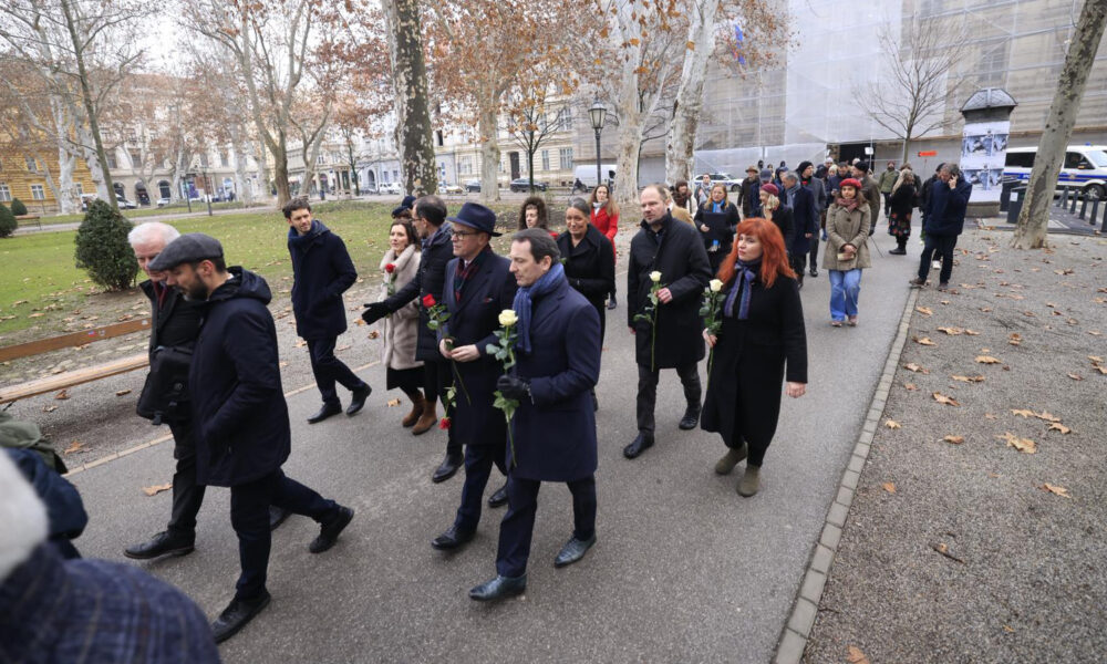 Povorka građana i predstavnika javnog života hoda kroz park u središtu Zagreba povodom Dana sjećanja na žrtve Holokausta. Sudionici u rukama drže bijele i crvene ruže, a atmosfera je dostojanstvena i tiha. Drveće bez lišća i pale smeđe lišće na tlu sugeriraju zimski ugođaj. U pozadini se vide zgrade s pročeljima i policijsko vozilo.