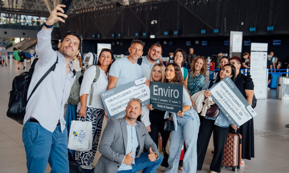 Grupa nasmiješenih putnika pozira za zajednički selfie u zračnoj luci. Drže natpise s porukama poput „I mi putujemo – Hrvatska i Slovenija bez prepreka za osobe s invaliditetom“ i „Enviro – Brežice & Zaprešić“. U pozadini se vide šalteri za prijavu i moderni interijer terminala. Atmosfera je vedra i inkluzivna.