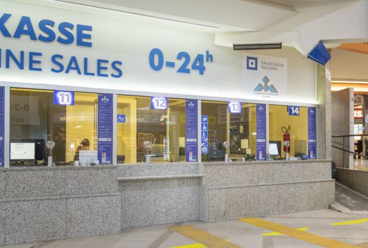 Fotografija prikazuje prodajno mjesto karata u glavnom autobusnom kolodvoru u Zagrebu. U prvom planu nalaze se šalteri označeni brojevima 11 do 14, smješteni unutar granitne strukture s prozorima od stakla. Iznad njih dominira veliki natpis "KASSE" i "LINE SALES 0–24h", što označava da su prodaja karata i linijske usluge dostupne 24 sata dnevno. Na gornjem dijelu konstrukcije vidljiv je logotip i naziv "ZAGREBAČKI HOLDING" te "AUTOBUSNI KOLODVOR ZAGREB", što potvrđuje lokaciju. Uz svaki šalter nalazi se plava informativna ploča s ikonama i tekstom, vjerojatno s detaljima o uslugama, vrstama karata ili linijama. Na podu se nalaze taktilne trake za osobe s oštećenjem vida, što ukazuje na prisutnost pristupačnih elemenata u prostoru. Cjelokupni ambijent djeluje funkcionalno, moderno i usmjeren na pružanje usluga putnicima u urbanom prometnom okruženju.