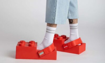 Na fotografiji je prikazan donji dio osobe koja nosi neobične crvene klompe u obliku LEGO kockica. Obuća izgleda masivno i geometrijski, s jasno izraženim ispupčenjima na vrhu koja podsjećaju na klasične LEGO čepove. Klompe su potpuno crvene, a sa strane imaju remen s vidljivim LEGO i Crocs logotipima. Osoba nosi bijele sportske čarape s tankim sivim prugama i svijetle, široke traperice skraćene iznad gležnjeva. Jedna noga je blago ispred druge, što dodatno naglašava neobičan oblik obuće. Pozadina je neutralna i svijetla, bez dodatnih elemenata, čime je fokus u potpunosti stavljen na klompe. Cijela fotografija djeluje minimalistički i studiozno, a kontrast između klasične odjeće i izrazito razigrane, gotovo karikaturalne obuće dodatno naglašava koncept suradnje LEGO-a i Crocsa.