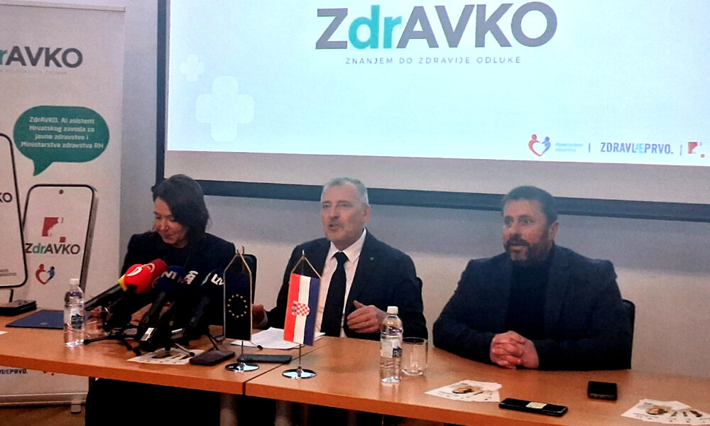 Troje govornika sjedi za stolom tijekom službenog predstavljanja digitalnog zdravstvenog asistenta ZdrAVKO. Ispred njih su mikrofoni, boce vode te zastavice Europske unije i Republike Hrvatske. U pozadini se nalazi veliki ekran s logotipom ZdrAVKO i sloganom „Znajem do zdravije odluke“.