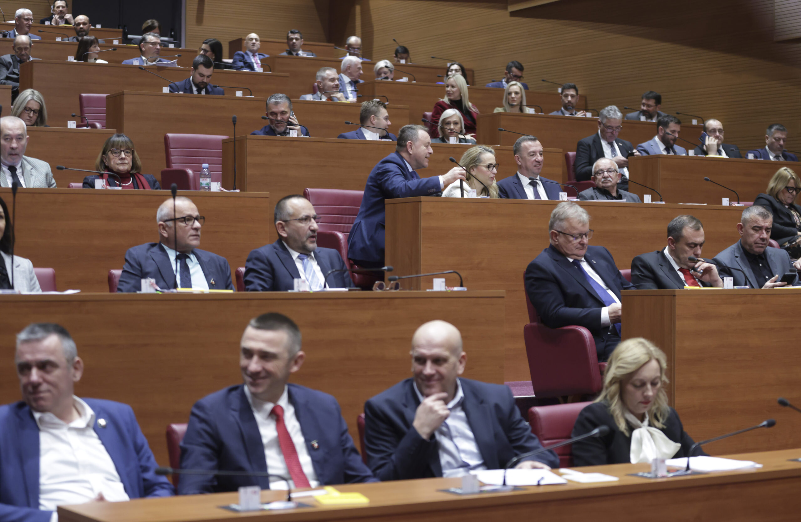 Zastupnici i zastupnice sjede u sabornici Hrvatskog sabora, raspoređeni u redove drvenih klupa s crvenim sjedalima. Dio njih razgovara, neki prate raspravu, a drugi pregledavaju dokumente ili mobitele. Prostor je osvijetljen i organiziran u polukružnim redovima, s mikrofonima ispred svakog mjesta, što upućuje na tijek saborske sjednice.