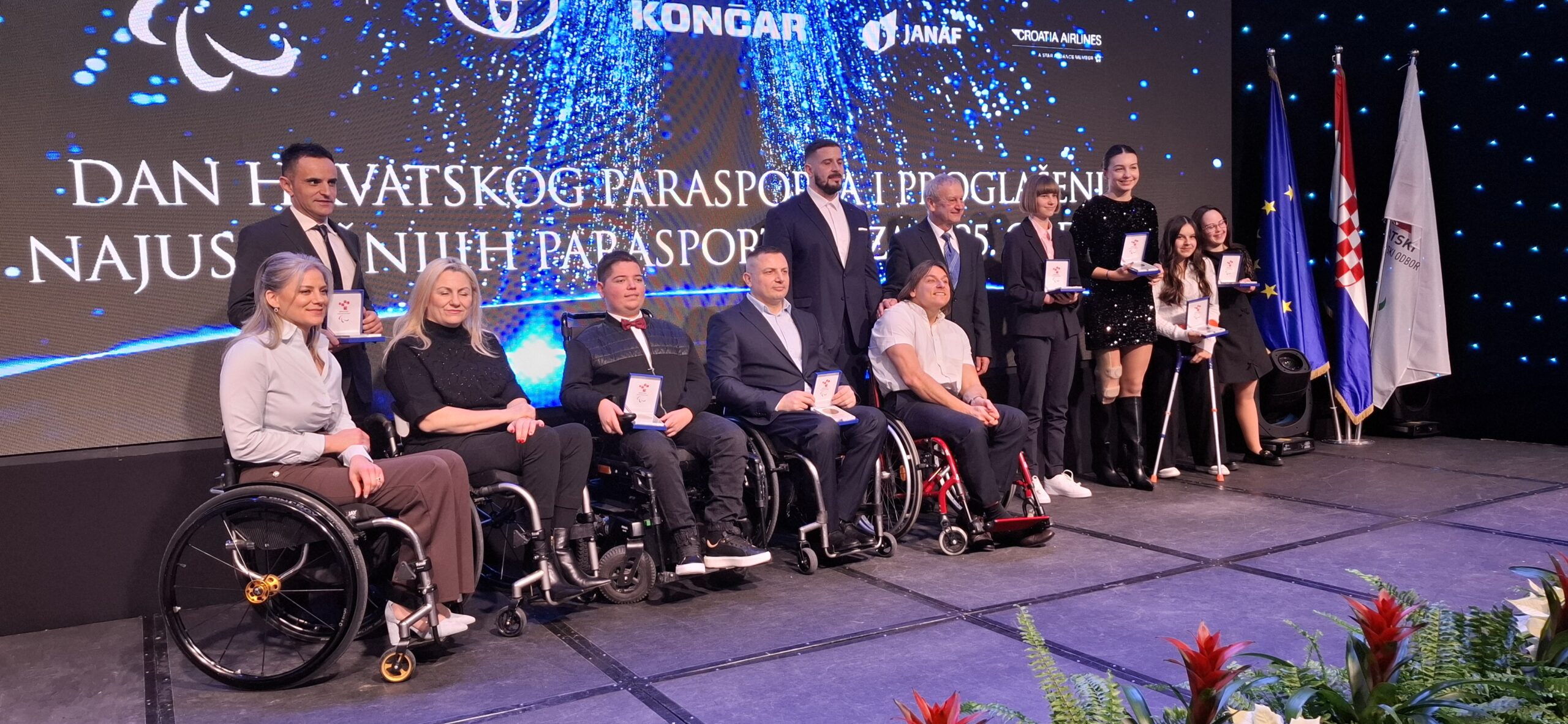 Fotografija prikazuje više ljudi na pozornici kako drže nagrade u ruci. Većina ih je u kolicima, dok neki stoje.