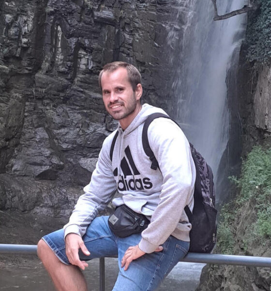 Muškarac u sivoj Adidas dukserici i trapericama sjedi na ogradi ispred visokog vodopada koji se spušta niz stijene. Na leđima nosi ruksak, a oko struka ima torbicu. Pozadina prikazuje stjenoviti kanjon s bujnom vegetacijom i tekućom vodom pri dnu.