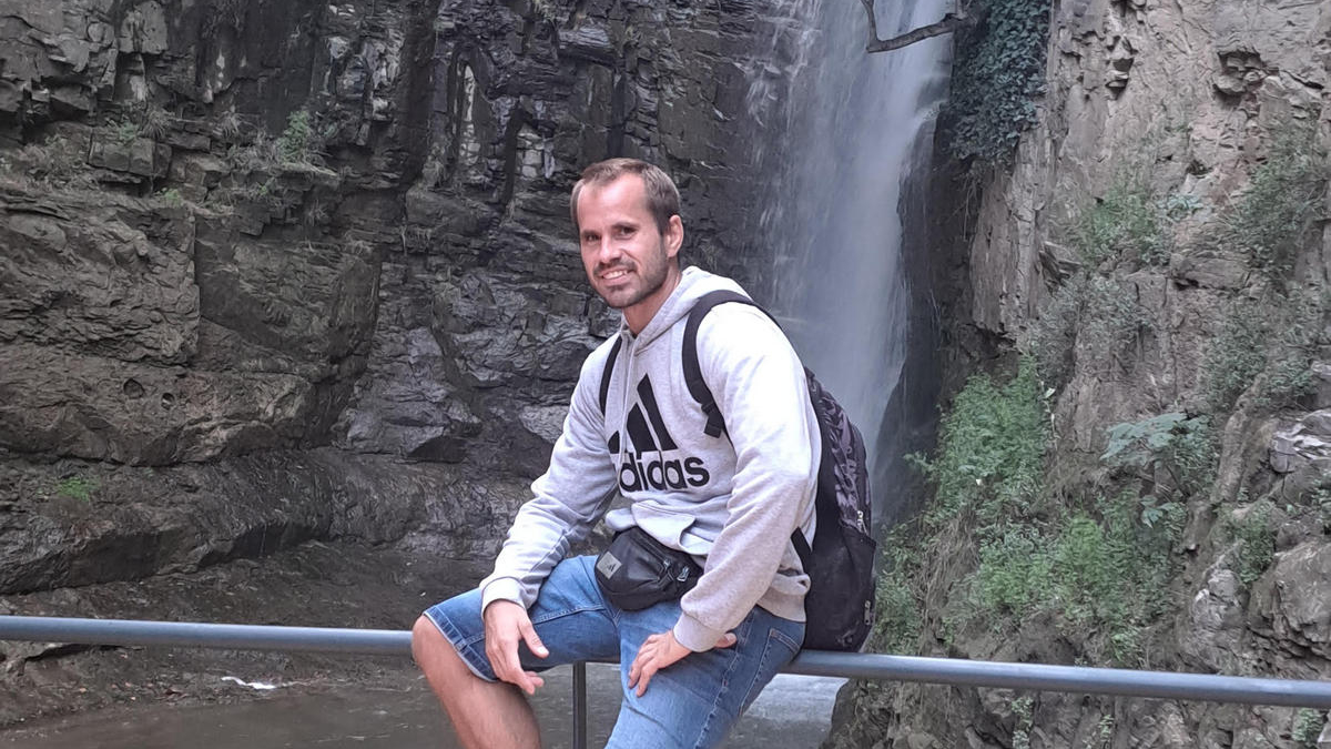 Muškarac u sivoj Adidas dukserici i trapericama sjedi na ogradi ispred visokog vodopada koji se spušta niz stijene. Na leđima nosi ruksak, a oko struka ima torbicu. Pozadina prikazuje stjenoviti kanjon s bujnom vegetacijom i tekućom vodom pri dnu.